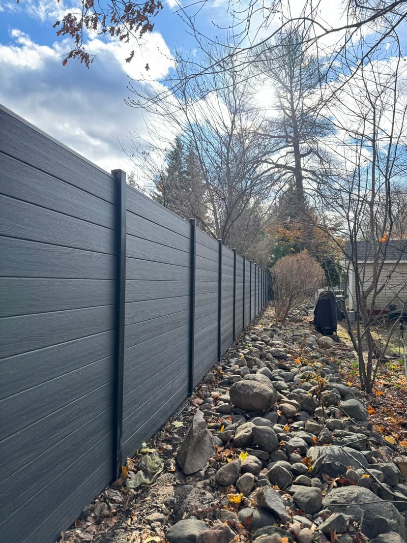  Composite charcoal fence along a rocky garden path with trees and cloudy sky in the background.
