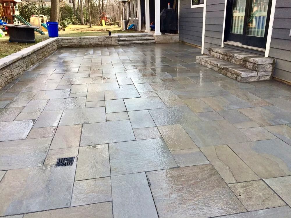 Beautiful stone patio with wet surface, surrounded by greenery and steps leading to a house.
