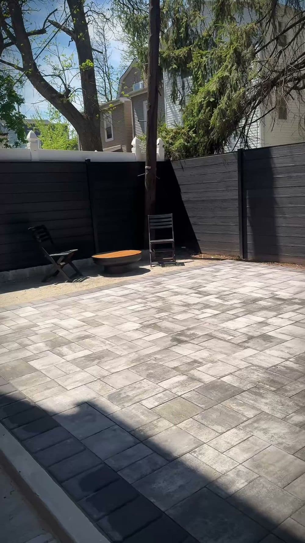Paved outdoor patio with black wooden fence, tree, and seating area.