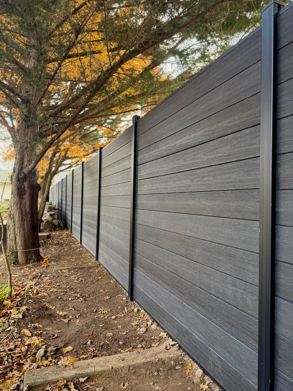 Modern dark wood fence alongside a tree in an autumn landscape with fallen leaves.