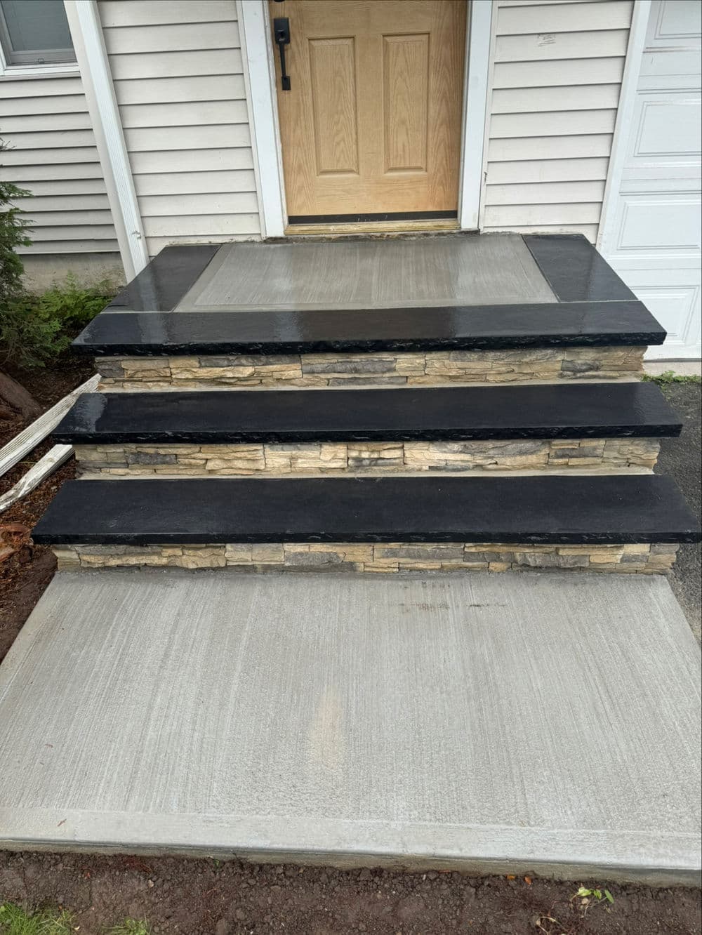 Three-step stone porch with black surfaces and concrete base leading to a front door.