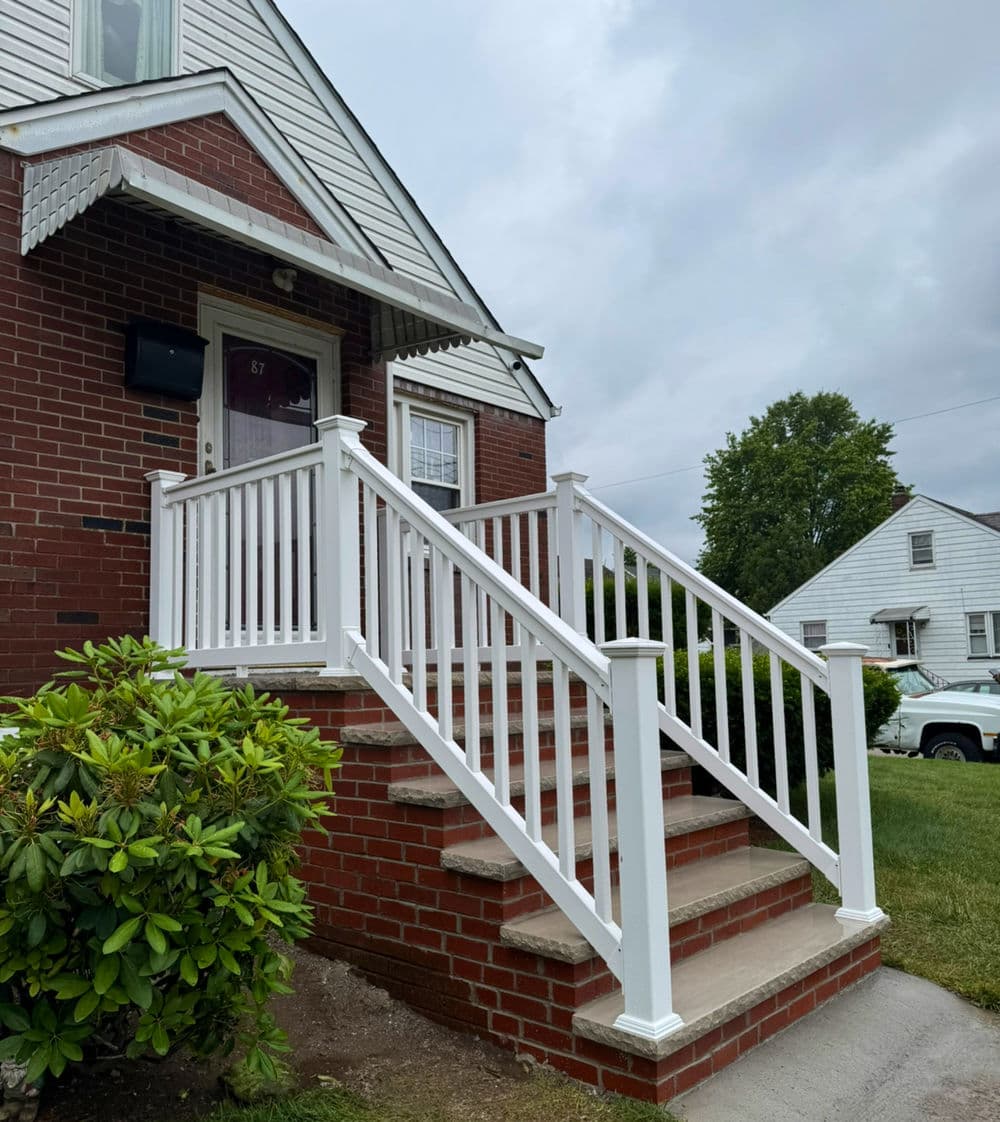 White railing on brick steps leading to a house entrance with green landscaping.