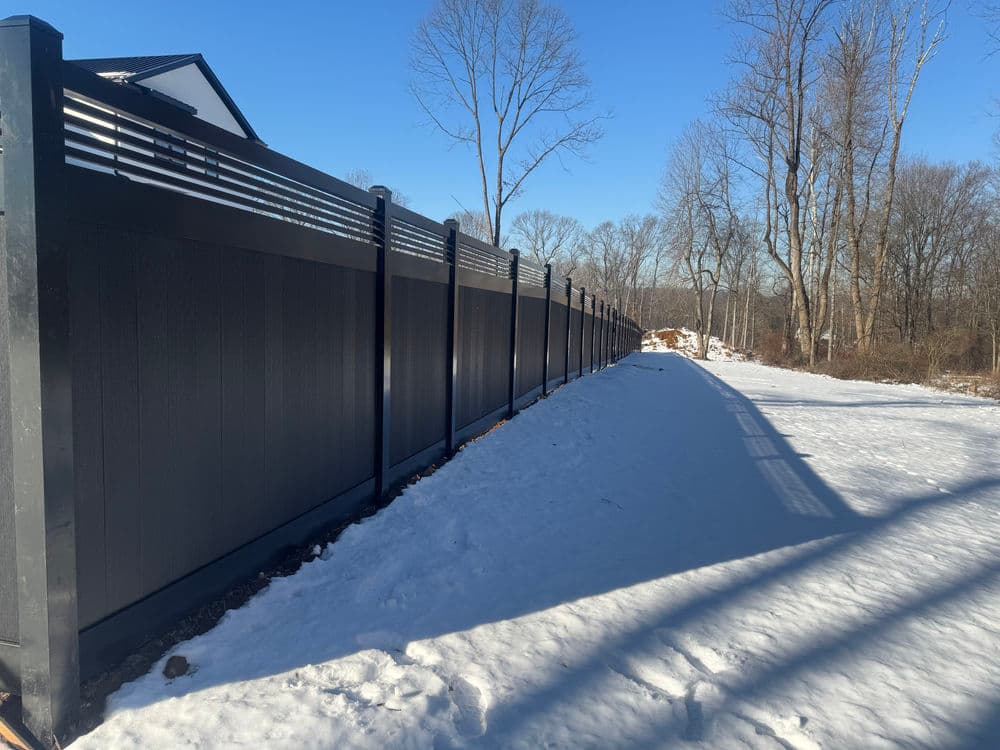 Charcoal composite fence along a snow-covered path in a winter landscape with bare trees.