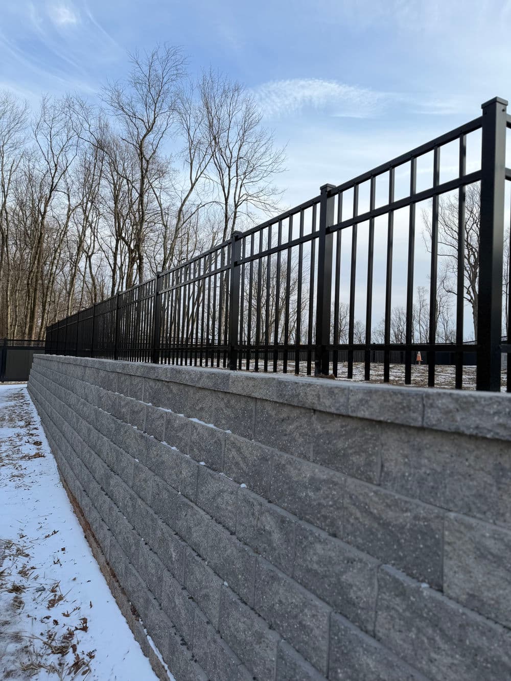 Black metal fence on a stone retaining wall with winter landscape in the background.