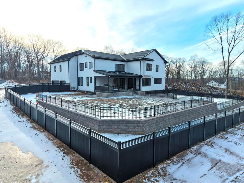 Modern two-story house with large patio, surrounded by a black composite fence and snowy landscape.