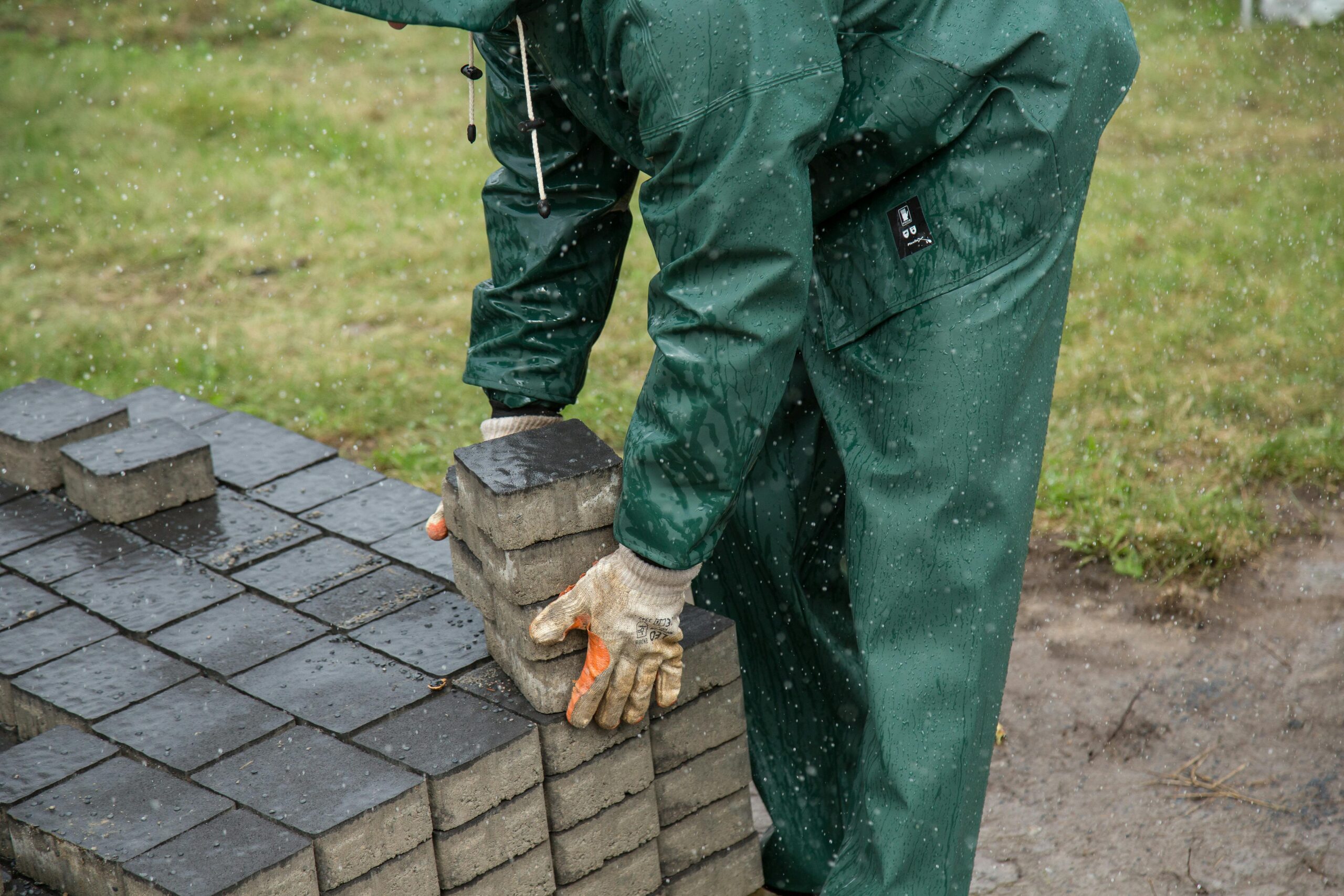 A worker in rain gear carefully places paving stones during a rainy day outdoor construction project.